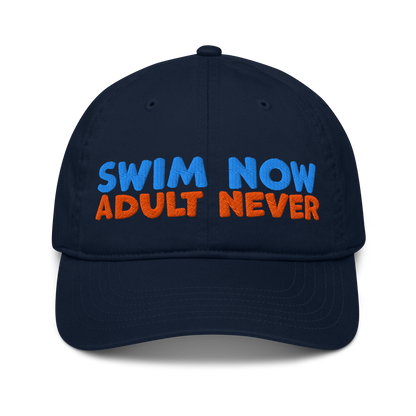 Navy blue organic cotton baseball cap with bold blue and orange embroidery reading “Swim Now, Adult Never.” Eco-friendly, vegan, and biodegradable.